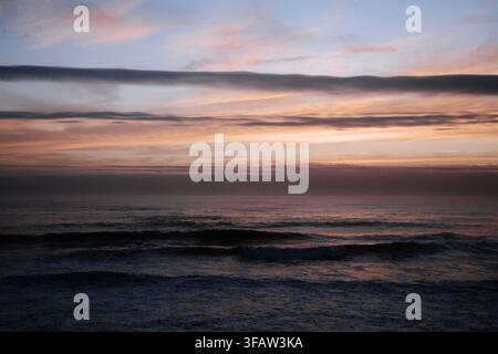 Un paysage côtier serein présentant des teintes douces rose, orange et violette se reflétant dans les nuages au-dessus d'un océan sombre et calme au crépuscule. Belle nature Banque D'Images