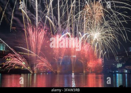 Un spectacle de feux d'artifice spectaculaire éclatant de lumières vives rouges, dorées et blanches au-dessus d'une rivière dans une ville européenne la nuit. Des réflexions brillantes scintillent Banque D'Images