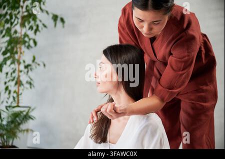 Massageuse faisant un massage thaï yoga. Femme thérapeute effectuant un traitement de massage traditionnel pour une patiente en utilisant le coude. Cliente assise, méditant, profitant d’un massage des mains dans une atmosphère calme. Banque D'Images