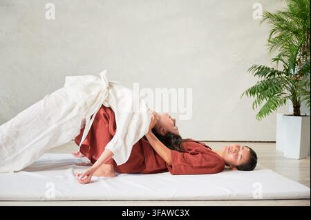 Masseuse faisant massage yoga thaïlandais. Thérapeute de femme faisant le traitement de massage traditionnel, étirant le corps de la patiente féminine. Client allongé sur tapis, profitant d'un massage des mains dans une atmosphère calme. Banque D'Images