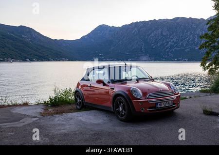 4 juillet 2026, Perast, Monténégro : Une Mini Cooper décapotable rouge garée sur une route côtière surplombant des eaux calmes de la baie. D’immenses chaînes de montagnes s’élèvent au-d Banque D'Images