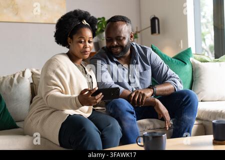 Couple afro-américain partageant l'écran d'un smartphone assis sur un canapé beige dans le salon Banque D'Images