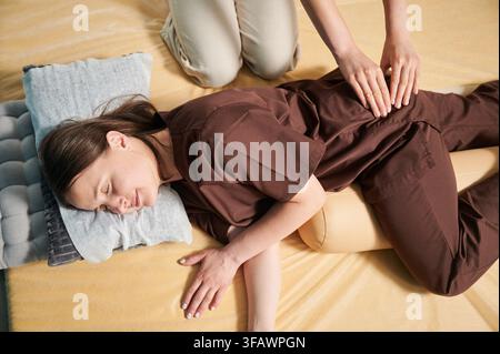 Masseuse faisant massage yoga thaïlandais. Vue rognée de thérapeute de femme faisant le traitement de massage traditionnel pour la patiente féminine. Client allongé sur tapis, méditant, profitant d'un massage des mains dans une atmosphère calme. Banque D'Images