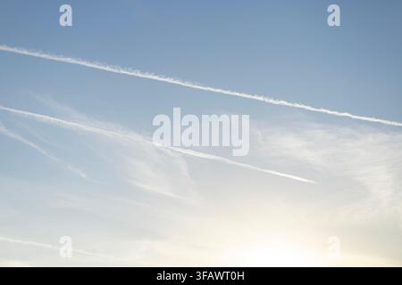 Des traînées de condensation blanc vif s'étendent sur un ciel bleu clair parsemé de nuages blancs et doux. Le soleil brille depuis le coin inférieur, créant une atmosphère très lumineuse Banque D'Images
