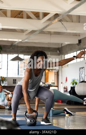 Classe de fitness afro-américaine effectuant un squat avec kettlebell de 5 kg, portant un haut gris sur un tapis dans le studio. Salle de sport, poids, groupe, mode de vie, industriel, motivation Banque D'Images