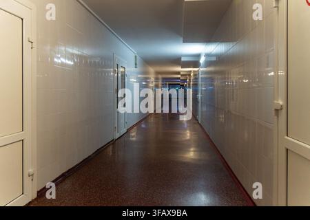 Couloir d'hôpital vide avec des murs carrelés blancs, des portes fermées, un éclairage fluorescent et un sol poli, créant un intérieur clinique propre avec de la profondeur Banque D'Images