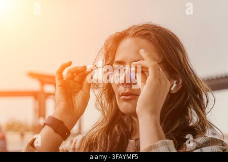 Femme lunettes coucher de soleil Jeune femme tenant des lunettes tout en portant des écouteurs sans fil et une montre intelligente en plein air à l'heure dorée Banque D'Images