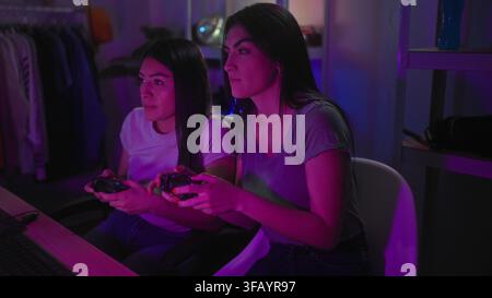Deux femmes tenant des manettes de jeu, les mains serrant les joysticks, souriant et se penchant en avant tout en jouant dans un studio avec des étagères néon et une lampe ; excitée Banque D'Images