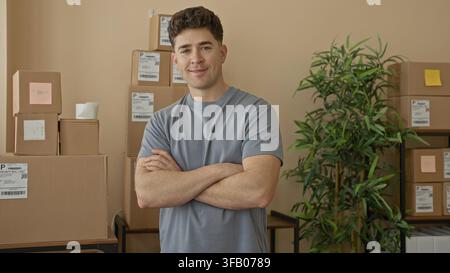 Homme aux bras croisés et cartons dans un petit bâtiment commercial, colis empilés sur des étagères et étiquettes d'expédition visibles ; entrepreneur confiant Banque D'Images