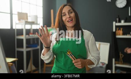 Peintre femme, âge moyen, en tablier vert tenant des pinceaux et pointant un pinceau vers un chevalet et une toile dans l'atelier ; créativité concentrée. Banque D'Images