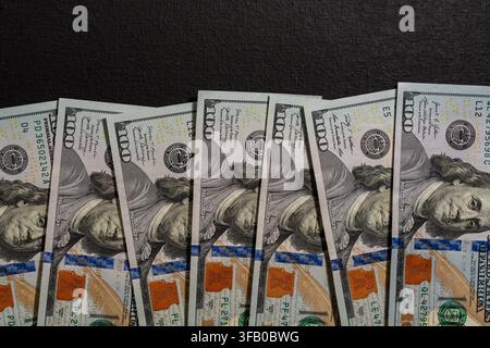 Billets de cent dollars américains alignés sur fond noir. Gros plan d'une prise de vue en contre-plongée de plusieurs billets de cent dollars disposés en rangée sur un texte noir foncé Banque D'Images