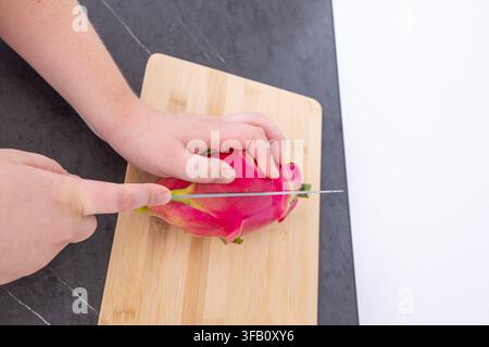 Vue rapprochée d'une personne coupant en deux une dragonne rose entière avec un couteau bien affûté, processus de préparation de la fruit pitaya tropicale biologique Banque D'Images