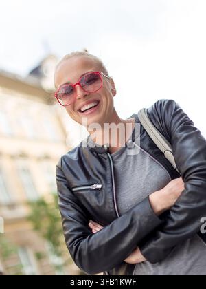 Femme souriante avec pPortrait d'une jeune femme heureuse et élégante dans une veste en cuir noire et des lunettes de soleil rouges teintées, riant les bras croisés en plein air par une journée lumineuse Banque D'Images
