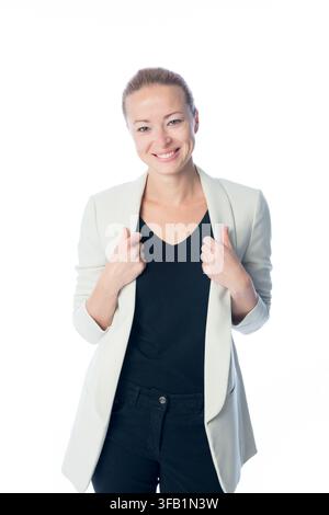 Portrait en studio d'une femme d'affaires blonde et joyeuse tenant les revers de son blazer gris clair, souriant chaleureusement à l'appareil devant un fond blanc uni Banque D'Images
