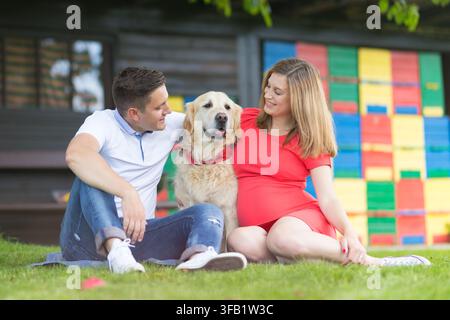 Jeun couple enceint heureux assis sur l'herbe dans un parc, souriant amoureusement à leur chien Golden Retriever positionné entre eux devant un fond coloré Banque D'Images