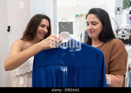 Une femme enceinte sourit en choisissant une robe bleue avec un employé du magasin dans une boutique de vêtements, mettant en avant la mode maternité, le shopping et le personnel Banque D'Images