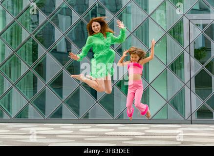 Mère et fille joyeuses sautent en l'air devant un bâtiment moderne en verre, portant des tenues vert vif et rose lors d'une journée estivale joyeuse dans le Banque D'Images