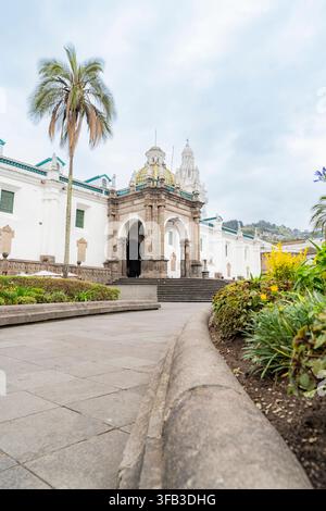 La cathédrale métropolitaine de Quito s'élève derrière un grand palmier, avec des dômes ornés, des murs coloniaux blancs, des arches de pierre, des marches de jardin et la ville en pente Banque D'Images