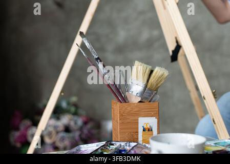 Gros plan de pinceaux à aquarelle, de couteaux à palette et de fournitures de peinture dans un porte-outils en bois à côté d'un chevalet, capturant l'atmosphère calme d'un artiste Banque D'Images