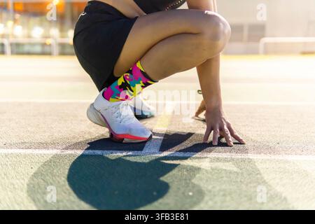 Close-up d'une coureuse accroupie à la ligne de départ sur une piste d'athlétisme en plein air, portant des vêtements de sport, des chaussettes colorées et des chaussures de course par temps chaud Banque D'Images