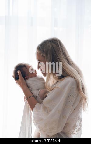 Mère tendre tient doucement son nouveau-né emmailloté près d'une fenêtre lumineuse, partage un regard aimant pendant les premiers jours à la maison et exprime la famille c Banque D'Images