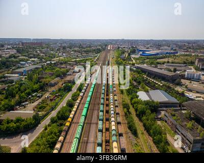 Un plan large par drone capture de longs trains de marchandises sur plusieurs lignes parallèles traversant un grand quartier industriel et un paysage de ville de banlieue sous le pont Banque D'Images