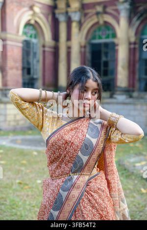 Une femme en sari traditionnel pose à l'extérieur avec un bras levé et la main posée sur la tête, debout devant un bâtiment orné de fenêtres en arc. Banque D'Images