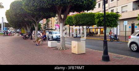 Promenade bordée d'arbres avec des gens sur des bancs, circulation routière et terrasses de café en plein air à Adra, Almería, Andalousie, Espagne, 14 août 2026. Banque D'Images