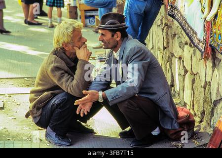 Deux hommes accroupis et discutant avec animation sur un marché de rue animé, scène de vie quotidienne spontanée. Tirana, Albanie, Europe Banque D'Images