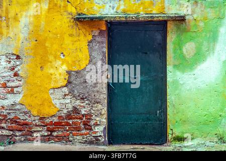 Mur érodé avec peinture jaune et verte écaillée, brique apparente et porte métallique vert foncé, texture de décadence urbaine. Albanie, Europe Banque D'Images