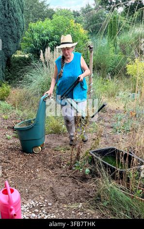 Une femme âgée travaille dans un jardin, portant un chapeau de paille, un haut bleu sans manches et un pantalon gris. Elle tient un genouillère de jardin et pousse une roue bleu-vert Banque D'Images