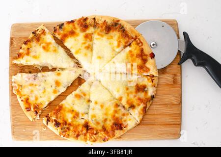 Une pizza au fromage est coupée en tranches avec un coupe-pizza reposant sur la planche en bois. Banque D'Images