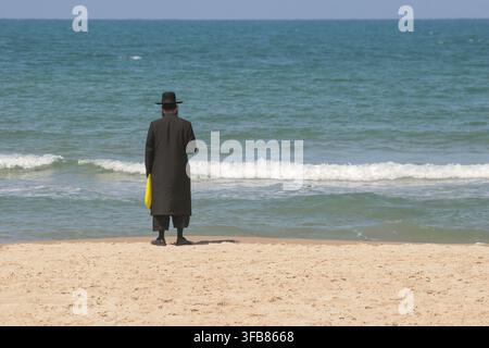 Homme juif religieux hassidique en vêtements noirs traditionnels debout sur une plage de sable avec vue sur la mer Méditerranée à Tel Aviv, Israël Banque D'Images