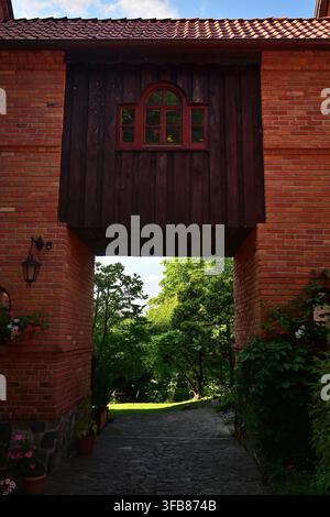 Une arche en brique avec une fenêtre en bois rouge sous laquelle passe un passage et une vue sur le chemin du jardin luxuriant. Banque D'Images