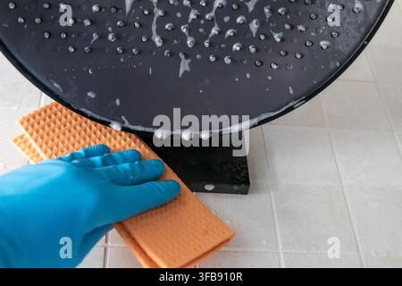 Une personne portant un gant bleu frotte un pommeau de douche avec une éponge orange texturée, créant de la mousse et des bulles sur la surface carrelée Banque D'Images
