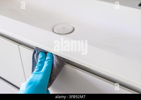 Une personne portant des gants de protection bleus essuie méticuleusement une surface de meuble de salle de bain blanc moderne avec un chiffon en microfibre gris doux, assurant la propreté Banque D'Images