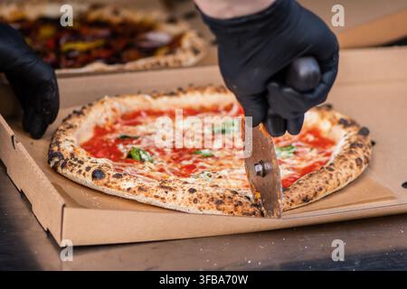 Faites pizzas avec des gants noirs utilisant un coupe-pizza roulant pour trancher une pizza Margherita de style napolitain fraîchement cuite dans une boîte à emporter en carton Banque D'Images