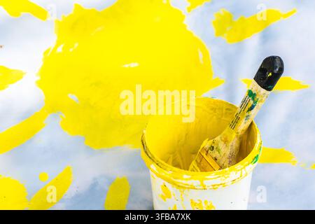 Petit seau rempli de peinture jaune vive et un pinceau reposant à l'intérieur, posé sur une surface blanche avec des éclaboussures de peinture jaune Banque D'Images