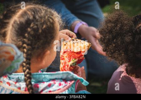 Enfant tenant une part de pizza avec sauce tomate, fromage fondu et mozzarella tout en étant assis en plein air avec la famille ou des amis Banque D'Images