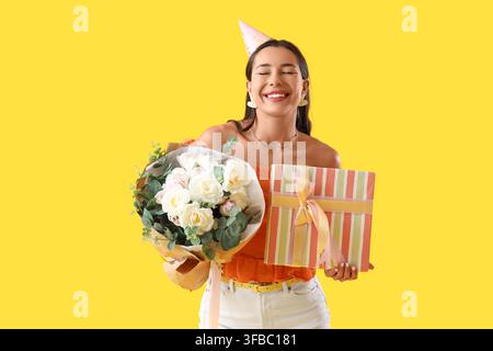 Célébration d'anniversaire et occasion spéciale, jeune femme heureuse avec chapeau de fête tenant des fleurs et une boîte cadeau Banque D'Images