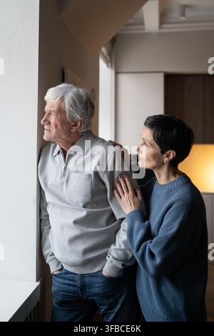 Un homme senior triste et réfléchi regarde par la fenêtre tandis que sa femme tend la main, consolant. Soutien familial, empathie Banque D'Images