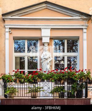 Façade historique d'immeuble avec une statue classique de cariatide entre les fenêtres, balcon en fer forgé et fleurs de géranium rouges dans la rue Lipska, Kiev. Banque D'Images