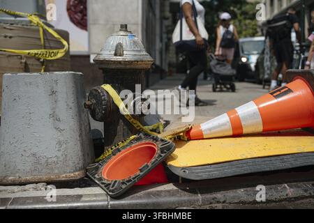 Un robinet d'incendie de rue est entouré de cônes de signalisation orange tombés et de ruban de sécurité sur un trottoir urbain animé. Les piétons passent devant une barrière de sécurité de chantier Banque D'Images