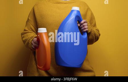 Femme dans un pull tricoté tenant des bouteilles en plastique orange et bleue contenant du gel de lavage liquide. Linge et tâches ménagères, fond jaune Banque D'Images