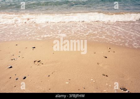 Des vagues douces s'écoulent sur une plage de sable chaud parsemée de petits galets avec une seule empreinte humaine visible au premier plan, Grèce. Banque D'Images