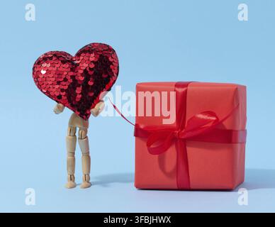 Un mannequin en bois tient un coeur rouge fait de paillettes et une boîte enveloppée de papier rouge, félicitations pour les vacances Banque D'Images