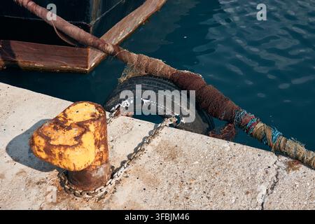 Vue rapprochée d'un bollard en métal rouillé et d'un épais câble d'amarrage enroulé d'un filet attaché à un quai en béton à côté d'un navire et d'une eau bleue, G Banque D'Images