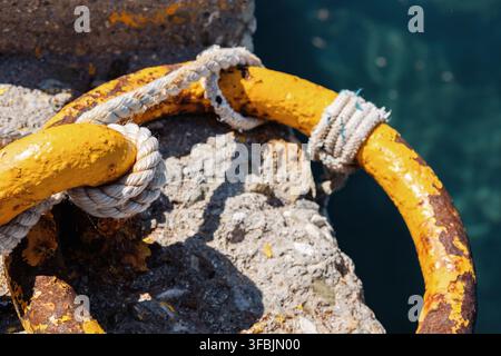 Gros plan d'un anneau d'amarrage en fer rouillé et jaune, solidement attaché avec une corde blanche épaisse sur un quai en béton patiné, à côté d'une eau de mer bleue en Grèce. Banque D'Images