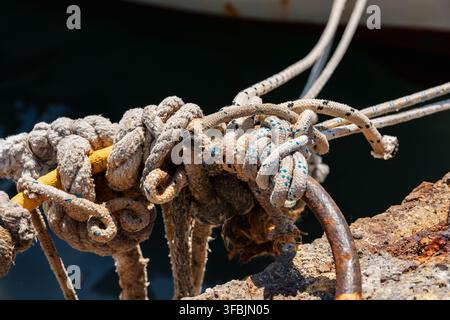 Gros plan détaillé de cordages nautiques usés et d'un épais cordage solidement attaché autour d'une bittes de mouillage métallique rouillée sur un quai en Grèce. Banque D'Images