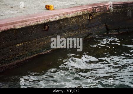Vue rapprochée d'un quai en béton usé avec des anneaux métalliques rouillés et un petit taquet le long du bord de l'eau sombre en Grèce. Banque D'Images